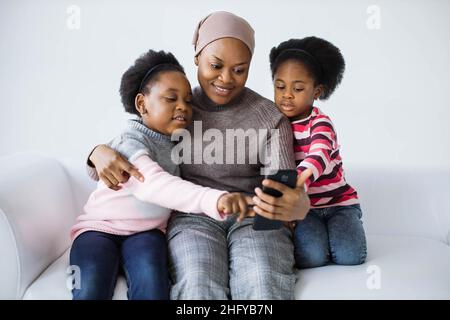Zwei niedliche Schwestern und ihre fürsorgliche Mutter verwenden ein modernes Smartphone, um Selfie zu machen, während sie sich auf der bequemen Couch ausruhen. afroamerikanische Familie, die Spaß hat, während sie Zeit zusammen verbringt. Stockfoto