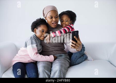 afroamerikanische Frau sitzt mit zwei hübschen Tochter auf der Couch und nimmt Selfie auf dem modernen Smartphone. Weißer Studiohintergrund. Familie und Gadgets Konzept. Stockfoto