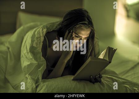 Die schöne Frau liest in ihrem Schlafzimmer vor dem Bett aufmerksam unter schwachem Licht ein Buch. Selbstentwicklung. Entspannung. Literatur. Stockfoto