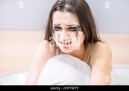 Eine Frau mit einem Gesicht, das von Mascara schmutzig ist, sitzt im Bett und weint. Finden Sie die schlechten Nachrichten heraus. Ärgern Sie sich. Leiden. Depression. Untröstlich Stockfoto