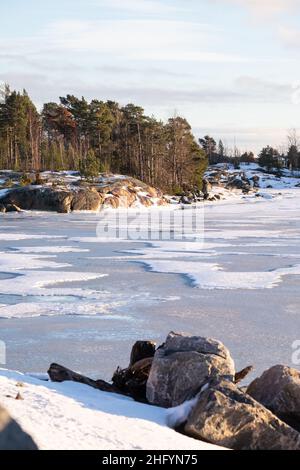 Helsinki / Finnland - 13. JANUAR 2022: Blick auf eine kleine Insel über einen gefrorenen Fluss an einem sonnigen Wintertag. Stockfoto