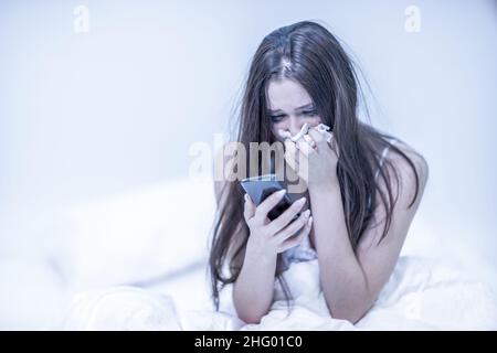 Eine Frau mit einem Gesicht, das von Mascara schmutzig ist, sitzt im Bett und weint, wischt sich mit einer Serviette und hält ein Telefon in den Händen. Depression. Holen Sie sich die schlechten Nachrichten. Stockfoto