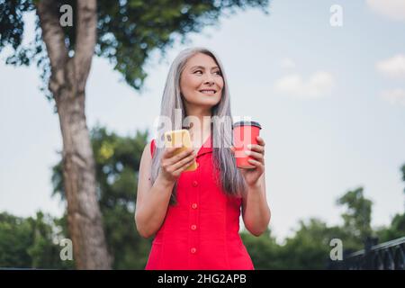 Foto von reifen attraktive asiatische Frau glücklich positives Lächeln Verwenden Sie Handy Internet online halten Tasse Kaffee im Freien zu Fuß Stockfoto