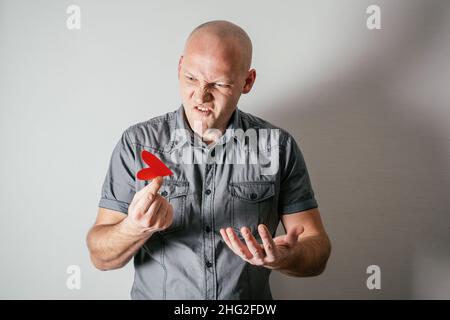 Ein gutaussehender, runzeliger Mann mittleren Alters in grauem Hemd hält das rote Papierherz mit zwei Fingern empört vor Hass auf weißem Hintergrund. Valentine's da Stockfoto