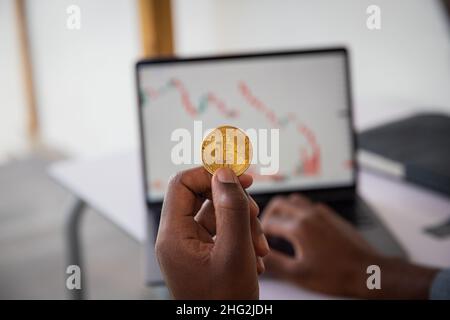 Nahaufnahme einer Bitcoin-Münze, die von einem Investor in der Hand gehalten wird, der den Wert der Kryptowährung mit dem Candlestick-Chart über seinen Laptop überprüft. Stockfoto