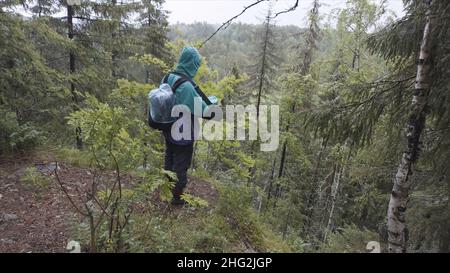 Männlicher Wanderer mit Reiserucksack, der während der Sommerwanderung am Rande des hohen Berges steht. Männlicher Erklärer, der auf einem Hügel steht und auf Baumkronen blickt. Stockfoto