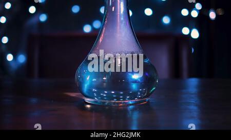 Die Kamera bewegt sich zur Glasvase für eine Shisha. Durchsichtige Flasche mit Wasser auf dunklem Hintergrund des Zimmers, dekoriert mit blauen Lampen. Stockfoto