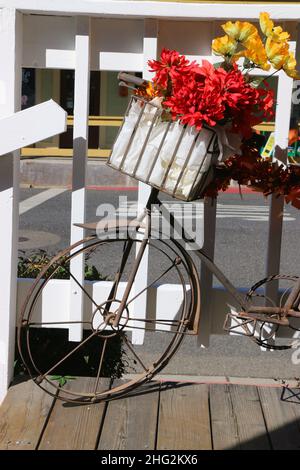 Vintage-Fahrrad mit leuchtend roten und gelben Seidenblumen in einem weißen Korbkorb steht vor einem Geschäft, einladend Herbst. Stockfoto