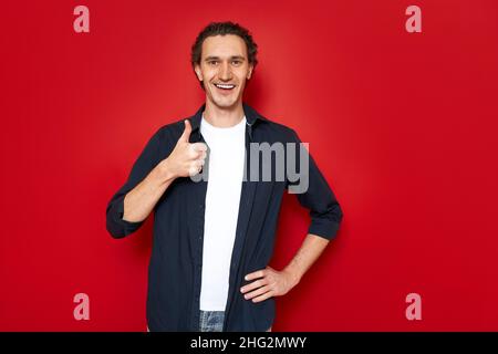 Fröhlich lächelnder Mann in blauem Hemd, weißes T-Shirt hält die Hand vor sich und zeigt seinen Daumen Stockfoto