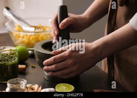 Frau, die Pesto-Sauce mit Mörser und Stößel auf dem Tisch in der Küche zubereitet Stockfoto