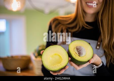 Porträt einer lächelnden jungen Verkäuferin auf der Schürze mit zwei Hälften frischer, reifer grüner Avocado Stockfoto