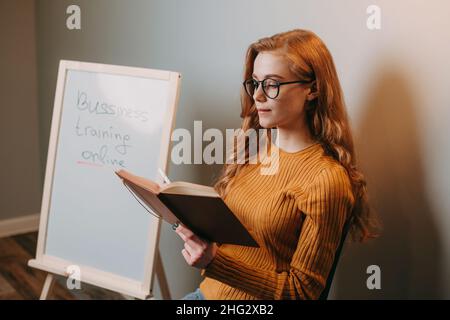 Foto eines Lehrers sitzt und hält ein Unterrichtsbuch in der Nähe des Whiteboards. Geschäftsfrau. Stockfoto