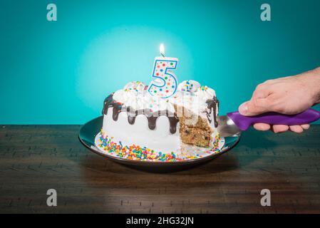 Eine Geburtstagstorte, die eine Scheibe fehlt, trägt eine Kerze in Form der Zahl 5, während eine Hand eine weitere Scheibe schneidet. Stockfoto