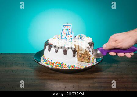 Eine Geburtstagstorte, die eine Scheibe fehlt, trägt eine Kerze in Form der Zahl 4, während eine Hand eine weitere Scheibe schneidet. Stockfoto