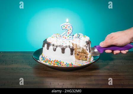 Eine Geburtstagstorte, die eine Scheibe fehlt, trägt eine Kerze in Form der Zahl 2, während eine Hand eine weitere Scheibe schneidet. Stockfoto