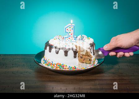 Eine Geburtstagstorte, die eine Scheibe fehlt, trägt eine Kerze in Form der Zahl 1, während eine Hand eine weitere Scheibe schneidet. Stockfoto