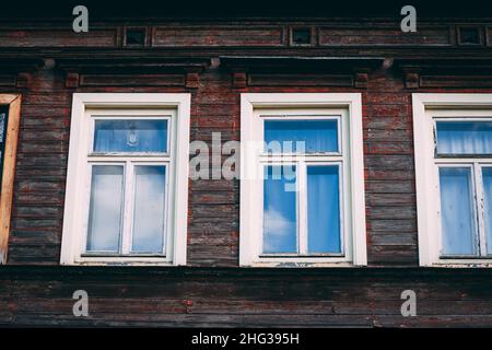 Die Wand eines alten Holzhauses mit roten Holzbrettern und weißen Fenstern an einem sonnigen Tag. Stockfoto