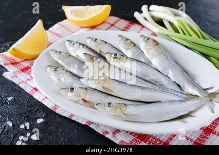 Frisches Meer kleiner Fisch auf einem Teller auf einem betonierten Hintergrund. Sardinen bereit zum Kochen mit Zitrone, grobem Meersalz und grünen Zwiebeln. Stockfoto