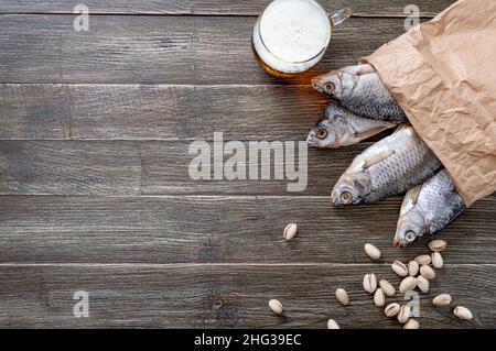 Gesalzener, getrockneter Fisch in einem Papierbeutel, frisches Bier, Pistazien auf einem Holztisch mit Platz für Kopien verstreut. Traditioneller Biersnack. Draufsicht, flach liegend Stockfoto