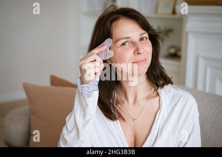 Süße kaukasische Frau mit Jade-Gouache-Schaber im Wohnzimmer auf dem Sofa im Bademantel, brünette Frau, die eine Gesichtsmassage mit einem Gouache-Schaber macht. Schönheits- und Körperpflege, Haut- und Gesichtspflegekonzept Stockfoto