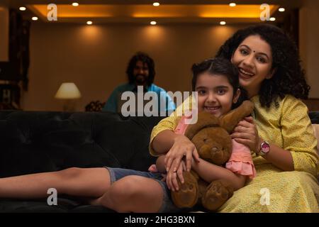 Mutter umarmte ihre Tochter auf dem Sofa und Vater beobachtete sie aus der Ferne Stockfoto