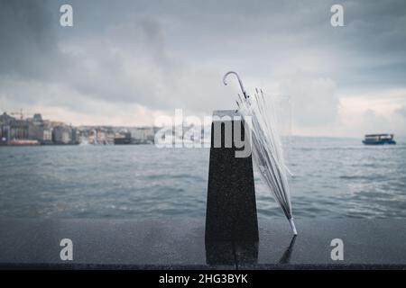 Vorderansicht eines geschlossenen transparenten Regenschirms, der an einem schwarzen Steinblock in der Nähe des Wassers mit Wolken oben und einer Küstenlinie im Hintergrund lehnt Stockfoto