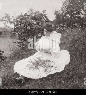 Vintage-Foto der Liebe. Er liebt mich, er liebt mich nicht. 1906 Frau saß auf dem Boden und pflückte Blütenblätter aus Blumen, während der Mann hinter dem Busch zuschaut. Stockfoto