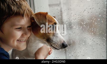 Kleiner Junge umarmt den Hund am Fenster. Freundschaft, Fürsorge, Glück, Neujahr Konzept. Stockfoto