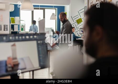 Ingenieur, der am Schreibtisch Baupläne auf einem Laptop mit Architekturmodell von Bürogebäuden ansieht. Architekt, der mit Computer arbeitet, um ein modernes Designkonzept für ein städtisches Wohnprojekt zu schaffen. Stockfoto