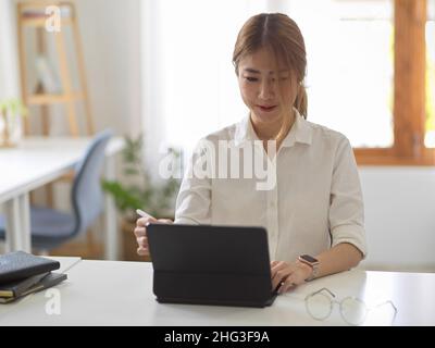 Eine schöne asiatische junge Geschäftsfrau oder Studentin, die an ihrem Arbeitsplatz einen digitalen Tablet-Computer verwendet. Stockfoto