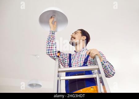 Glücklicher Elektriker, der auf einer Leiter steht und die LED-Glühbirne in die Deckenlampe schraubt Stockfoto