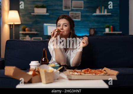 Frau nach der Büroarbeit, die einen Bissen aus einem Stück Pizza mit Heißlieferung nahm, während sie am Tisch Fernsehen und ein Fast-Food-Menü zum Mitnehmen hatte. Person, die auf der Couch im Wohnzimmer sitzt und zum Abendessen zum Mitnehmen isst. Stockfoto