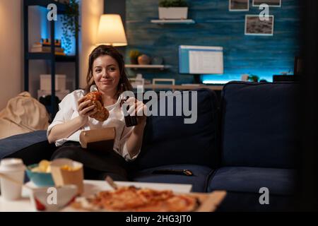 Glückliche Frau, die einen leckeren Hamburger zum Mitnehmen mit einer Bierflasche auf der Couch zum Entspannen und Fernsehen im Familiensitz hält. Lächelnde Person, die Burger zum Mitnehmen und ein Fast Food-Liefermenü hat. Stockfoto