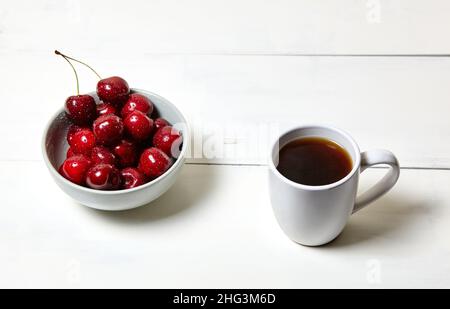 Weiße Tasse mit Kaffee und süßen Kirschen in einer Keramikschale auf weißem Holzhintergrund, Nahaufnahme. Leckeres Frühstück, morgendliches Routinekonzept Stockfoto