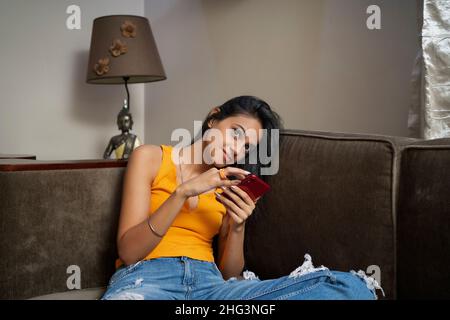 Eine junge Frau, die alleine in ihrem Zimmer sitzt und ihr Mobiltelefon benutzt, um etwas zu denken. Stockfoto
