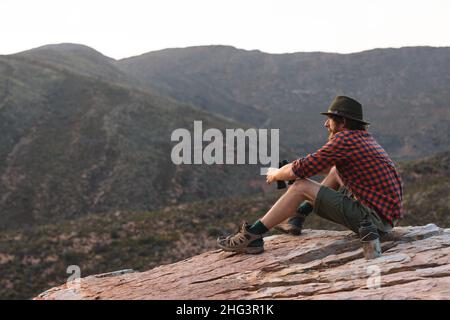 Ganzjährige Seitenansicht eines jungen Abenteurers, der mit einem Fernglas auf dem Gipfel sitzt und die Berge betrachtet Stockfoto