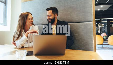 Junge Profis lächeln sich an einem modernen Arbeitsplatz an. Zwei glückliche junge Geschäftsleute, die zusammen an einem Laptop arbeiten. Zwei erfolgreiche junge ent Stockfoto