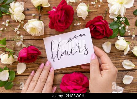 Hände mit handgeschriebenen KARTENBLUMEN, umgeben von roten und cremefarbenen Rosen, schließen sich auf einem Holztisch an. Femminine romantische Liebeserklärung nahe Blumen, Stockfoto