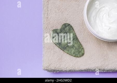 Gesichtspflege und Massage. Gesicht Gua Sha Stein mit Creme und auf einem Handtuch auf lila Hintergrund Stockfoto
