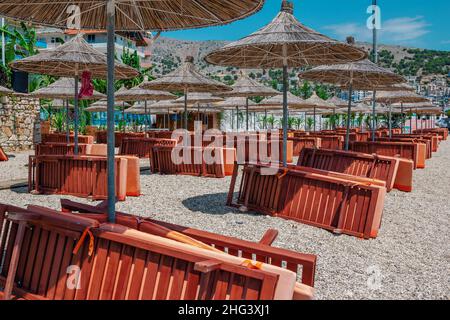 Blick auf den leeren Strand – Holzliegen und Schattenschirme in der Nähe des Meerwassers, der Stadtgebäude und der Berge am Horizont. Landschaft von verlassenen Sommer re Stockfoto