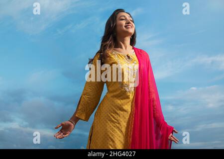 Glückliche Frau in gelbem Salwar, die unter dem blauen Himmel mit ausgestreckten Armen steht Stockfoto