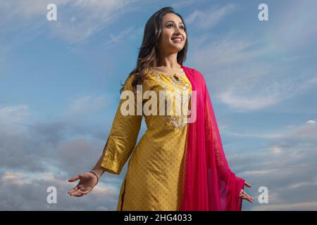 Glückliche Frau in gelbem Salwar, die unter dem blauen Himmel mit ausgestreckten Armen steht Stockfoto