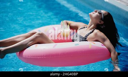 Erfreut Frau in Sonnenbrille hält Glas mit Cocktail und Schwimmen auf aufblasbaren Ring im Pool Stockfoto