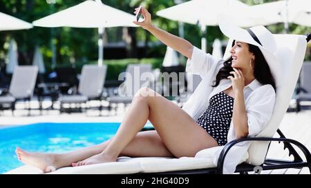Lächelnde Frau im Strohhut beim Selfie, während sie sich auf einer Liege in der Nähe des Swimmingpools ausruhte Stockfoto