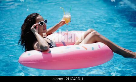 Erfreut Frau Anpassung Sonnenbrille, während Glas mit Cocktail und Schwimmen auf aufblasbaren Ring im Pool Stockfoto