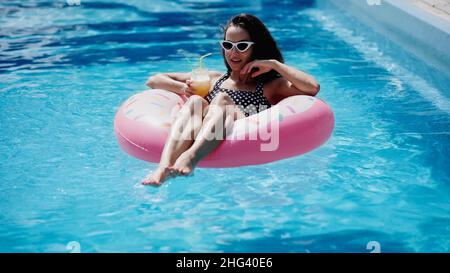 Brünette Frau in stilvollen Sonnenbrille hält Glas mit Cocktail und Schwimmen auf aufblasbaren Ring im Pool Stockfoto