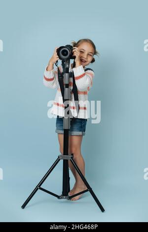 Fröhliches kleines Mädchen in gestreiftem Pullover, das hinter einer Digitalkamera auf einem Stativ auf blauem Hintergrund steht. Sie schaut auf die Kamera. Stockfoto