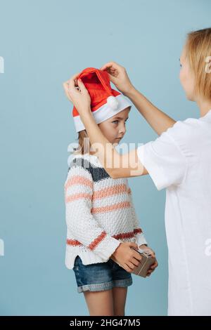 Mama festlegt weihnachtsmütze für ein widerstrebendes kleines Mädchen, das eine Geschenkbox in ihren Händen über blauen Hintergrund hält. Stockfoto