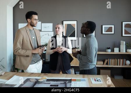 Drei kreative Designer diskutieren Arbeitspunkte, während sie am Arbeitsplatz stehen und in der Pause Kaffee trinken Stockfoto