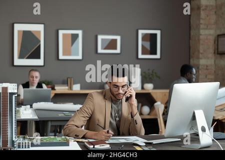 Junger, moderner Geschäftsmann, der sich ein Bild von der Inneneinrichtung zeichnet und mit dem Kunden am Mobiltelefon spricht, während er am Arbeitsplatz sitzt Stockfoto